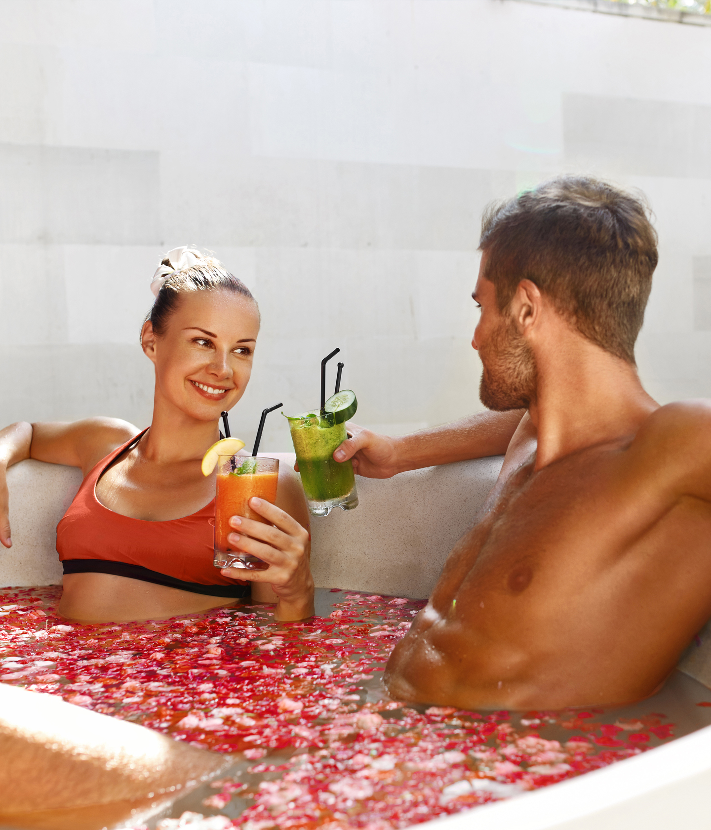 Ein Paar in Badekleidung entspannt sich in einem mit Blumen gefüllten Whirlpool, jeder hält einen Cocktail in der Hand und lächelt sich an.